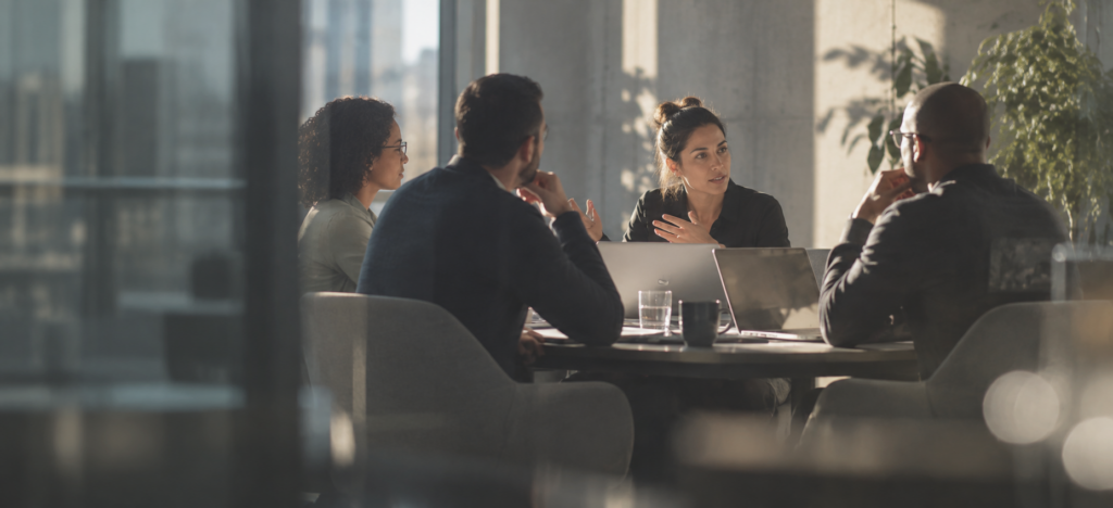 Four colleagues discussing ideas around a conference table in a sunlit modern office.